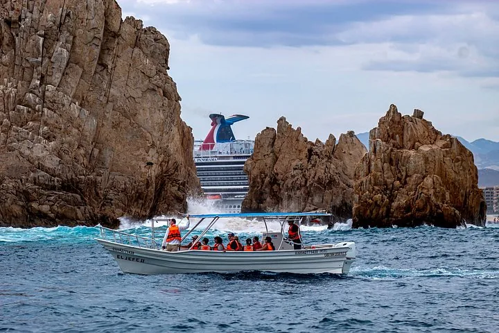 Un petit bateau transporte des passagers près de falaises rocheuses, avec un grand navire de croisière en arrière-plan.