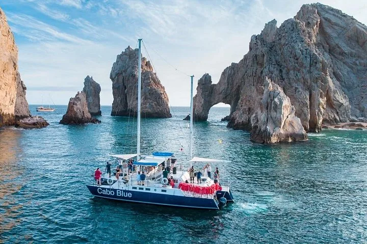 A catamaran carries passengers near large rock formations, including an arch, in the ocean.