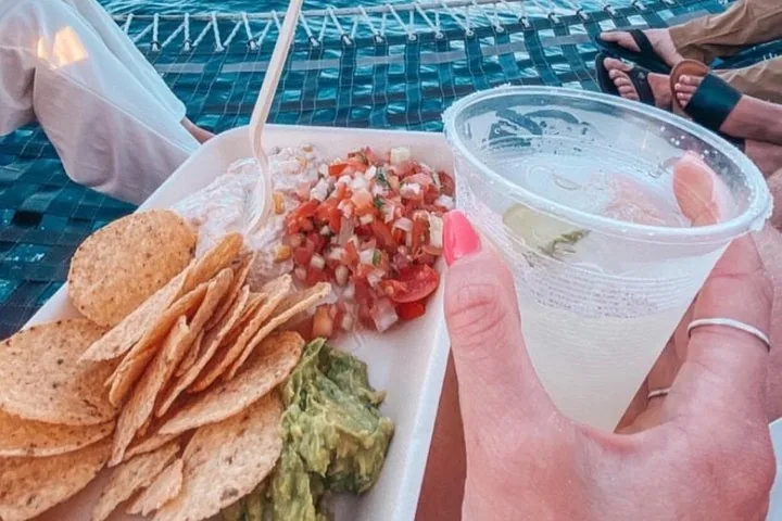Une main tient une boisson dans un gobelet en plastique à côté d'une assiette de chips, de salsa et de guacamole.