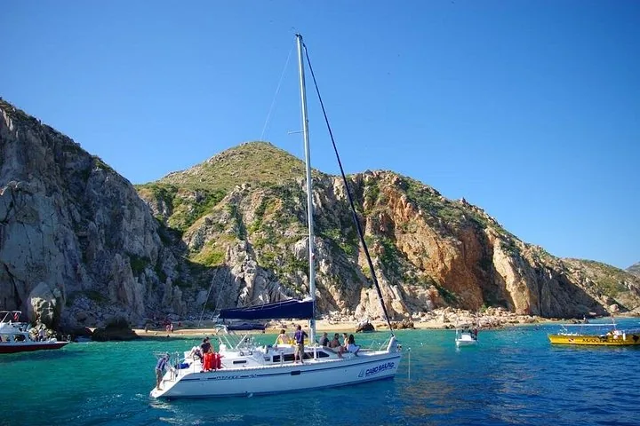 A sailboat anchors near a beach with rocky hills in the background.